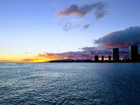 Beautiful sunset over the ocean at Ala Moana Beach Park in Honolulu, Hawaii. The photo shows the orange and blue sky with scattered clouds, the deep blue ocean with small waves, and the skyline of Honolulu with tall buildings in the background.