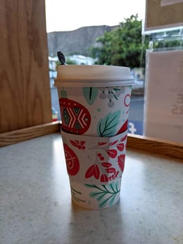 Honolulu - December 8, 2021: To-go cup of coffee with a floral design and a straw on a counter. The photo shows a window with a view of a building and trees in the background. 