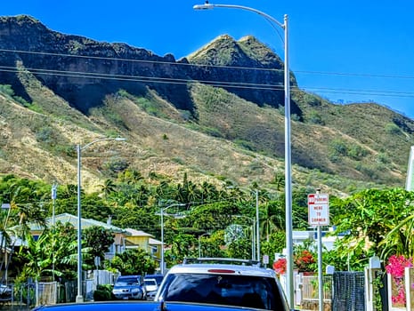 Honolulu - Juyne 14, 2023:  Diamond Head, a volcanic tuff cone on the Hawaiian island of Oʻahu, seen from a street in the Kapahulu neighborhood. The photo shows a black car, a street lamp, a no parking sign, and power lines in the foreground. The sky is blue with white clouds and the palm trees and houses are in the middle ground.