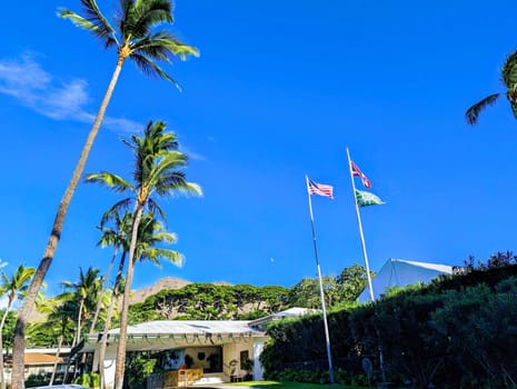 Honolulu - November 22, 2023:  A view of the Pacific Club Oahu with American and Hawaiian flags flying in the breeze. The photo shows the white building with a green roof and a covered patio. The sky is a bright blue with a few wispy clouds. The grassy area has palm trees and other tropical foliage.