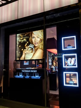Waikiki - December 7, 2017:  Victoria’s Secret store window display at night in Waikiki, Hawaii. The display features a large poster of a woman wearing a black bra and holding a perfume bottle, and several smaller screens with images of lingerie and perfume. The window is framed in black and has a pink striped awning. 