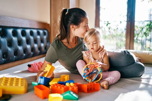 Mom kisses on the head a little girl playing with a puzzle rattle sitting on the bed. High quality photo