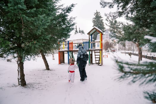 Mother and a small child walk holding hands to a snowy playground among the trees. Back view. High quality photo