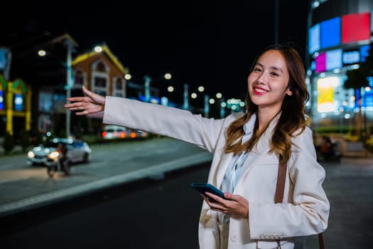 Asian businesswoman standing hail waving hand taxi on road in busy city street at night, beautiful woman smiling using mobile phone application hailing with hand up calling cab after late work