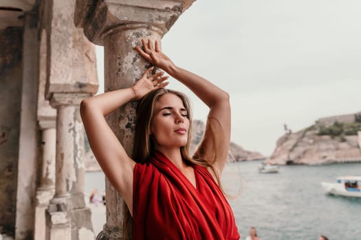 Side view a Young beautiful sensual woman in a red long dress posing on a volcanic rock high above the sea during sunset. Girl on the nature on overcast sky background. Fashion photo