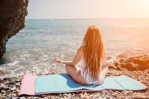 Young woman in swimsuit with long hair practicing stretching outdoors on yoga mat by the sea on a sunny day. Women's yoga fitness pilates routine. Healthy lifestyle, harmony and meditation concept.