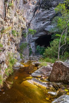 Secluded stream flows into a vast cave amidst craggy rocks and verdant flora.