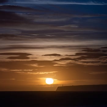 "Silhouetted lighthouse on cliffs against a dusky, cloud-streaked sky above a calm sea, creating a peaceful scene."