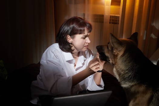 Adult mature woman with big shepherd dog in white shirt in dark room. Concept of loneliness and love for animals, pets