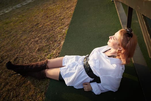 Beautiful girl with red hair in white shirt in open wooden pavillion in village or small town. Young slender woman and sky with clouds on background on autumn, spring or summer day