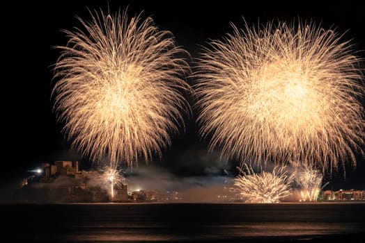 A night photograph showcasing Peniscola village with the castle, houses, and golden palm-style fireworks illuminating both the sky and the base