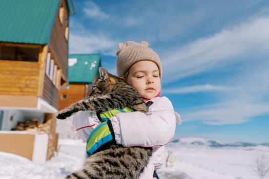 Little girl hugs a tabby cat while standing in the snow near a wooden cottage. High quality photo