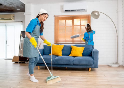 Wide shot of Asian housemaid or housekeeper team help to clean living room together with one use broom clean floor and other use feather duster for cleaning window.