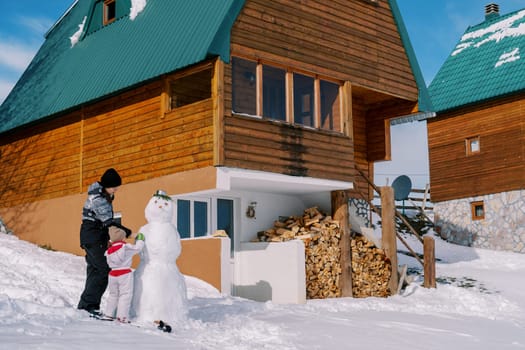 Mom and little girl decorate a snowman near a woodpile of a wooden cottage in the snow. High quality photo