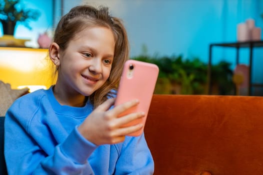 Young child girl texting share messages content on smartphone social media applications online, watching relax movie. Happy teenager kid uses mobile phone at home in evening night room sits on sofa