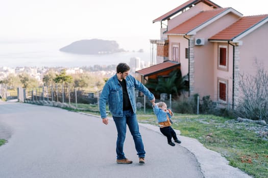 Dad is circling a little girl holding her hand on the road on a slope in a village above the sea. High quality photo