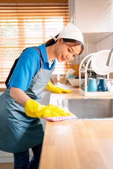 Vertical image of Asian housekeeper or housemaid woman use towel to clean area in kitchen near sink with day light and she look happy to work in house.