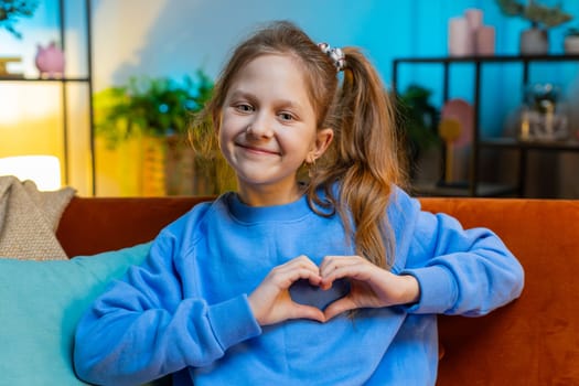 I love you. Child kid girl makes symbol of love, showing heart sign to camera, express romantic feelings, express sincere positive feelings. Charity, gratitude, donation. Female teen at night home