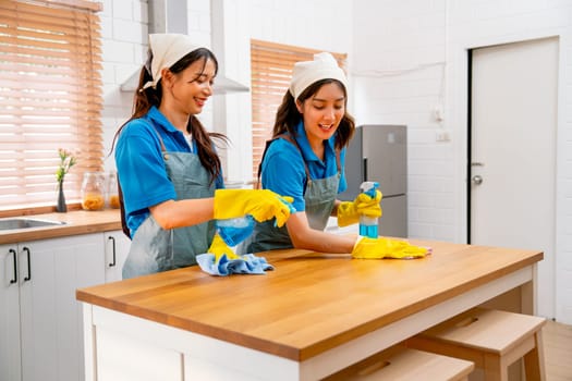 Two Asian housekeeper or housemaid women enjoy to work and teasing each other with one woman spray cleaner to top surface during clean table in kitchen at customer house.