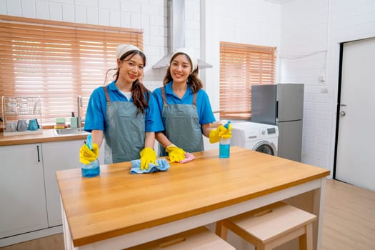 Portrait of two Asian housekeeper or housemaid women stand together near table in kitchen and hold towel for cleaning and look at camera with smiling.