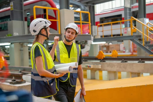 Side view of train factory workers man and woman discuss about work in workplace of maintenance center with electric train on rail is in the background.