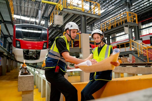 Wide shot of engineer worker woman sit and hold drawing paper to discuss with technician man in front of electric train in factory workplace or maintenance center.