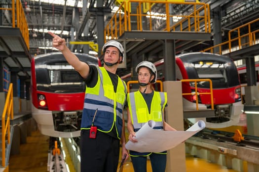 Two professional engineer or technician workers man and woman discuss with drawing paper and the man point to left side also stay in front of electric train in factory workplace or maintenance center.