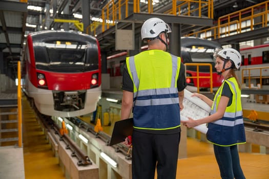 Back of technician or engineer workers man and woman discuss together with drawing paper during work in front of electric train in factory workplace or maintenance center.