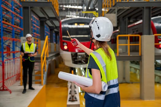 Back of technician or engineer worker woman use walkie talkie point to her co-worker during work in front of electric train in factory workplace or maintenance center.