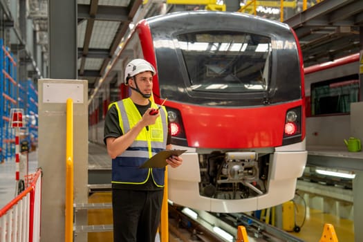 Technician or engineer worker man hold walkie talkie to contact with team member or staff also stay in front of electric train in factory workplace or maintenance center.