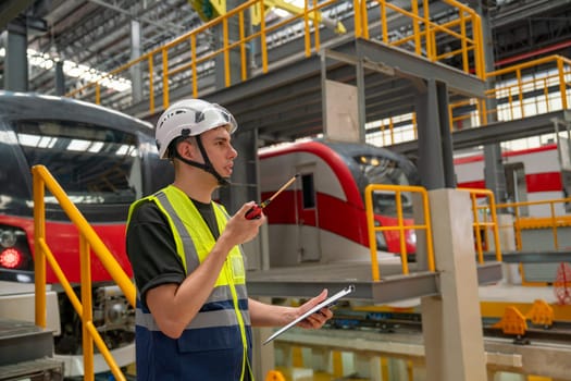 Technician or engineer worker man hold walkie talkie to contact with team member or staff also stay in front of electric train in factory workplace or maintenance center.