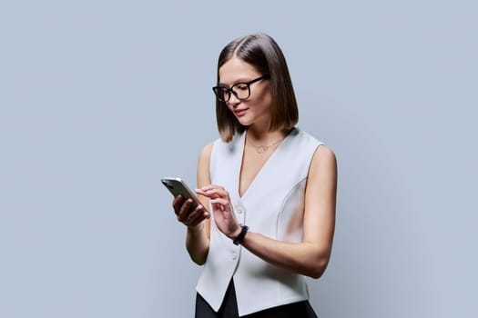 Young trendy business woman in glasses using smartphone on gray background. Smiling female texting chatting. Technology mobile applications online internet business work communication study education