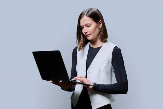 Portrait of young business fashionable woman using laptop on gray studio background, serious female looking at computer screen. Work, study, business education technology concept