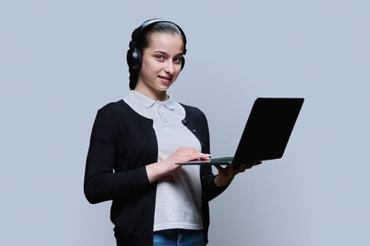 Teen girl student wearing headphones using laptop, looking at camera on grey studio background. Audio video internet technologies in education learning, online lessons, adolescence high school concept