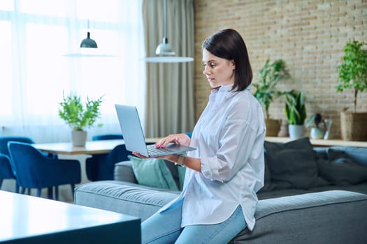 Young relaxed woman sitting on sofa at home in living room, using laptop for work, freelancing, learning, blogging, communicating, typing on keyboard