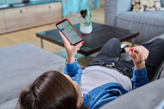 Young woman lying at home on sofa in living room using smartphone for online communication, chatting with friends, colleagues, family. People, technology, lifestyle concept