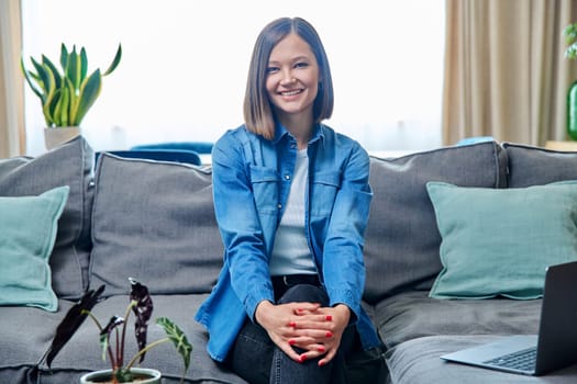 Portrait of young smiling attractive woman looking at camera, sitting on sofa in living room at home. Lifestyle, leisure, youth concept