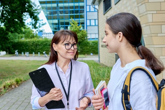 Teenage girl high school student with backpack talking to female teacher, mentor, coach, standing outdoors on educational building background. Adolescence, education, knowledge, communication