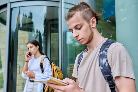 Hipster guy teenager student 18, 19 years old with backpack notebooks standing outdoor, educational building background. Youth, education, lifestyle concept