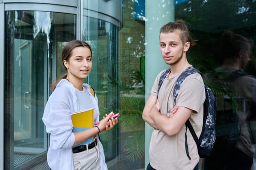 Portrait of two teenage friends, guy and girl with backpacks looking at camera, outdoor, educational building background. Friendship, communication, education, high school college concept