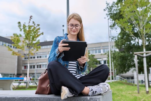 Girl student teenager outdoor near school building. Smiling teenage female with backpack, looking at screen of digital tablet. Adolescence, education, learning concept