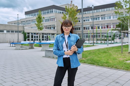 Middle-aged confident woman school teacher, mentor, pedagogue, psychologist, counselor, social worker with digital tablet in hands posing near school building, outdoor. Education, teaching, school