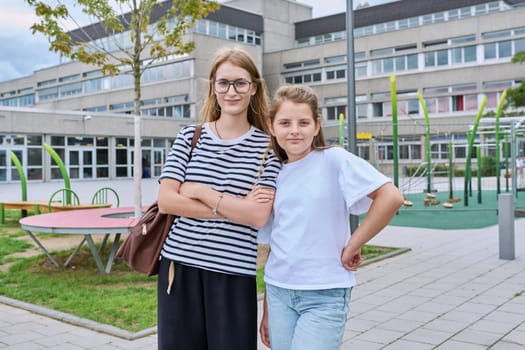 Portrait of two junior and high school schoolgirl sisters posing together near the school building. Adolescence, childhood, family, friendship, education, learning concept