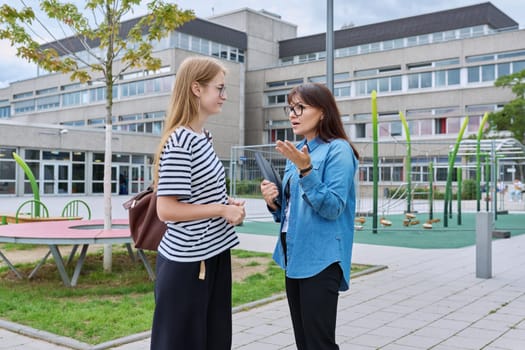 Talking female teacher and teenage high schoolgirl outdoor, school building background. Meeting communication student girl with backpack and mentor counselor. Education, adolescence, learning concept