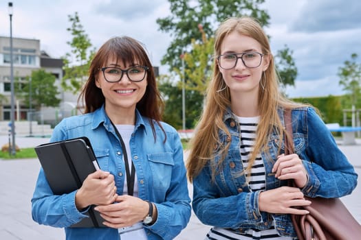 Teacher and teenage schoolgirl looking at camera together outdoor, school building background. Meeting communication student girl with backpack and mentor counselor. Education, adolescence, learning concept