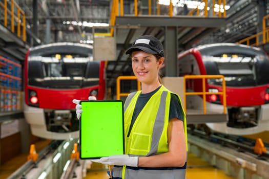 Pretty engineer or technician worker woman hold tablet with green screen and look at camera with smiling also stay in front of electric train in factory workplace or maintenance center.