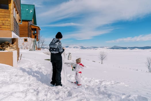 Little girl stands with a carrot near her mother making a snowman near the cottage. High quality photo