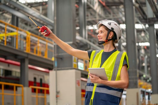 Pretty factory worker woman point to left side using walkie talkie and also hold tablet with smiling in concept of happiness to work in workplace of industrial center.