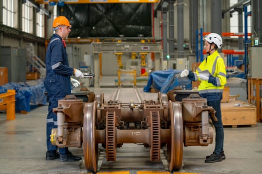 Factory workers man and woman discuss about work and stand in workplace with wheel and some parts of electric train are in front of them.