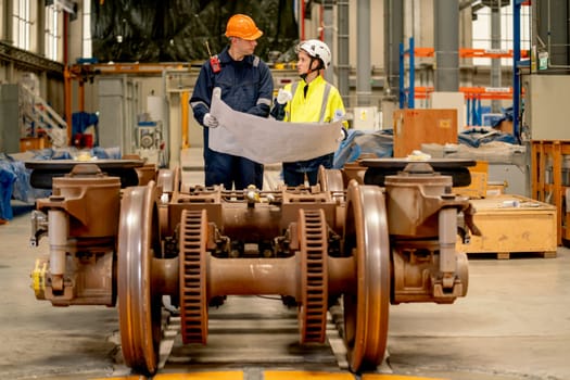 Professional factory workers man and woman discuss about the project using drawing paper or plan in workplace with wheel and some parts of electric train are in front of them.